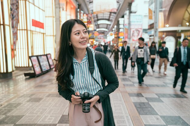 woman enjoy the view of the shopping center and holding camera ready to photograph. young traveler walking in the indoor street. happy tourist having fun on summer travel jp.