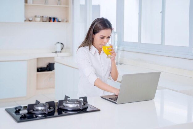 Woman enjoying juice while using laptop in kitchen