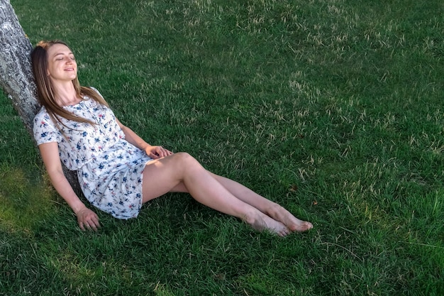 Woman enjoying and relaxing sitting on the lawn leaning her back on a tree on a summer day, she smiles with closed eyes with pleasure