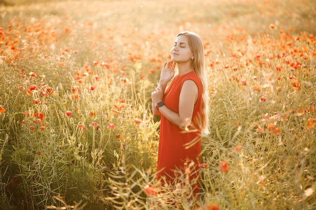 Женщина европейской внешности с длинными светлыми волосами и в красном летнем платье стоит на цветущем маковом поле.