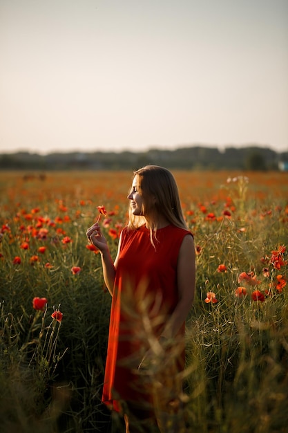 A woman of European appearance with long blonde hair and a red summer dress, she stands in a blooming poppy field