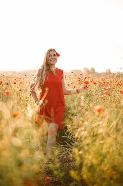 Una donna dall'aspetto europeo con lunghi capelli biondi e un vestito estivo rosso, sta in piedi in un campo di papaveri in fiore Foto Premium
