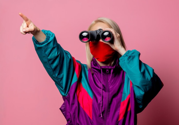 Woman in face mask and 80s clothes with binoculars on pink