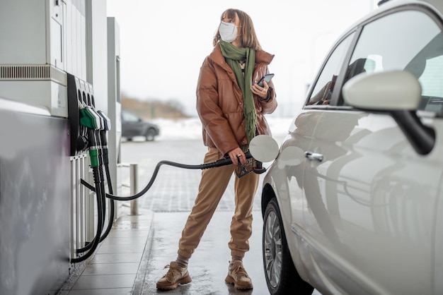 Woman in face mask refueling car with a gasoline using smart phone