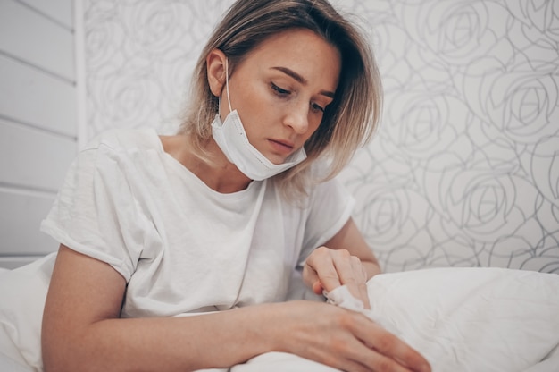 Женщина в защитной маске для лица, лежа в постели во время изоляции от коронавируса, домашний карантин чистит руки дезинфицирующим средством для рук, используя ватку со спиртом, чтобы вытереть, чтобы избежать загрязнения COVID-19