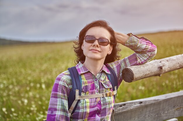 暗い眼鏡をかけた女性農民は、家畜のために牧草地の木製の柵にもたれかかって、夏の日に太陽に向きを変えました。