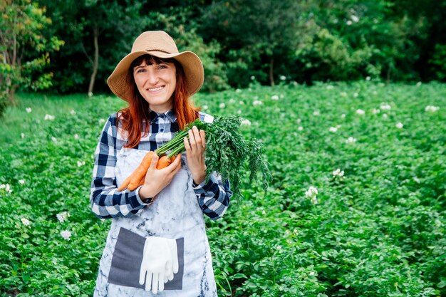 Woman farmer holds a bunch of carrots in a straw hat on background of a vegetable garden