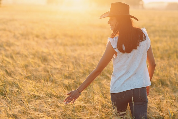 写真 カウボーイの帽子をかぶった女性農夫が夕暮れの農作物の小麦畑で耳に手をつけて歩いています