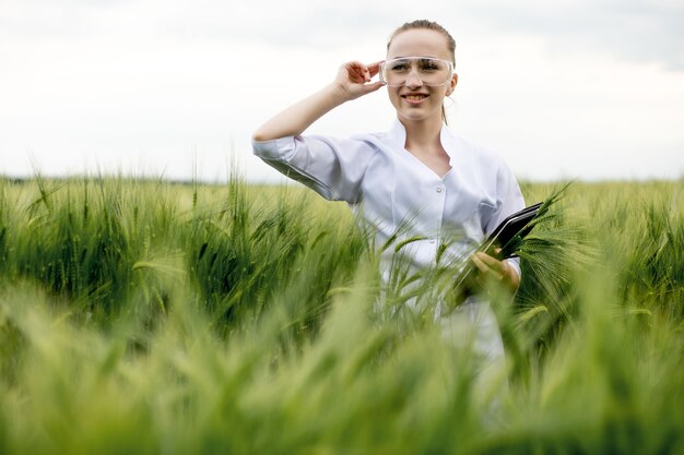 woman farmer wearing white bathrobe is checking harvest progress on tablet at the green wheat field