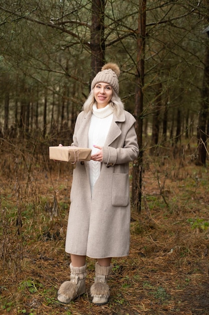 Woman in fashion warm clothes holding gift box in Kraft paper packaging on nature background