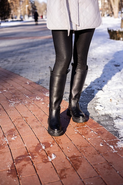 Woman in fashionable black boots on white snow close up Womens legs in stylish winter leather boots