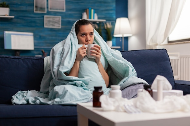 Photo woman feeling sick in blanket with warm drink at home having seasonal virus symptoms. unwell young adult in pain headache resting on couch with medical treatment against coronavirus