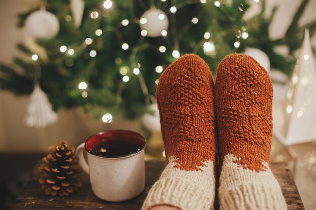 Woman feet in cozy woolen socks and cup of warm tea on\
background of christmas tree in lights