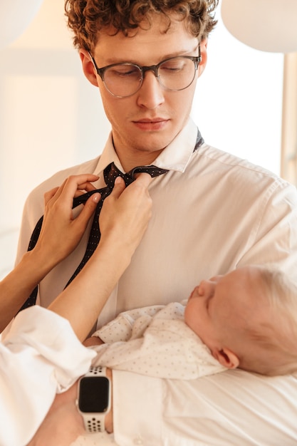 Woman fixing husbands tie while he is holding their baby son, standing at home, getting ready for work