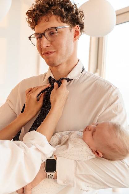 Woman fixing smiling husbands tie while he is holding their baby son, standing at home, getting ready for work