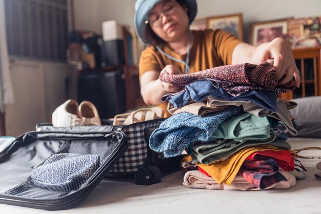 女性が選んだ持ち物の山から服を折りたたんで、ベッドの上の荷物に入れて休暇を過ごすためのパッキング服に焦点を当てる