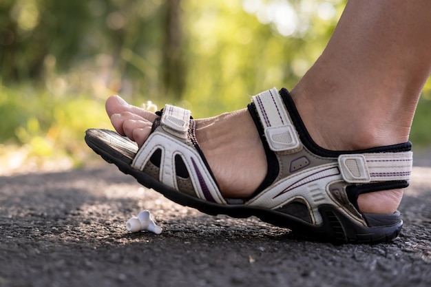 サンダルを履いた女性の足が、屋外のアスファルトの歩道にある失われたワイヤレスヘッドフォンを踏む