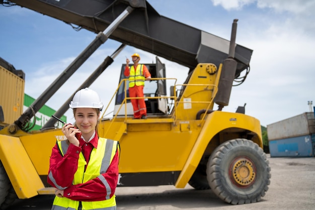 Woman foreman smile using walkie talkie in front of cargo\
forklift talking with driver in forklift manager in white helmet\
safety supervisor in container custom terminal port concept import\
export