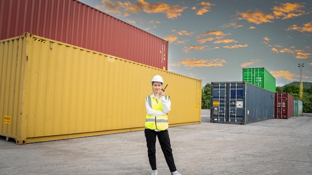 Woman foreman smile using walkie talkie in front of cargo\
forklift in warehouse manager in white helmet safety supervisor in\
container custom terminal port concept import export