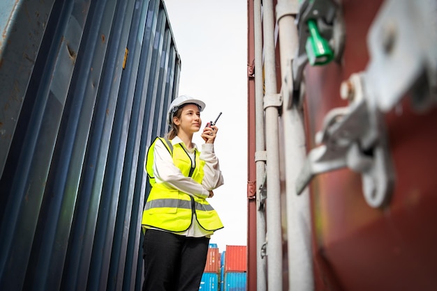 Woman foreman smile using walkie talkie in front of cargo\
forklift in warehouse manager in white helmet safety supervisor in\
container custom terminal port concept import export