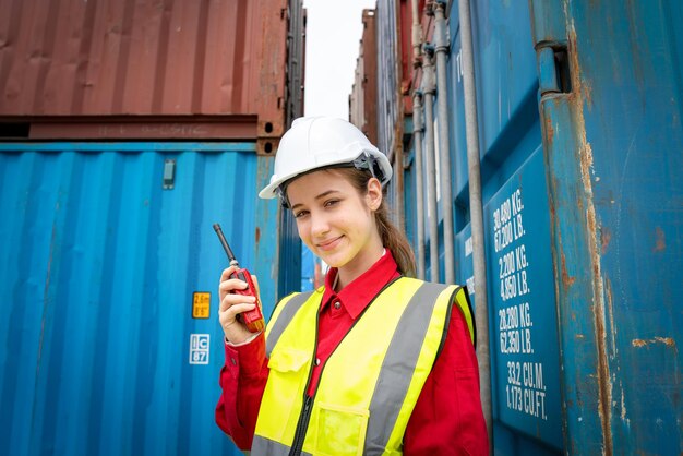 Woman foreman smile using walkie talkie in front of cargo\
forklift in warehouse manager in white helmet safety supervisor in\
container custom terminal port concept import export