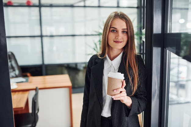 Woman in formal clothes standing indoors in the office with cup of drink.