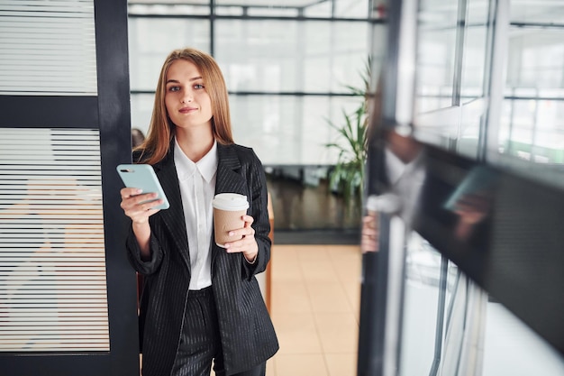 Woman in formal clothes standing indoors in the office with cup of drink.