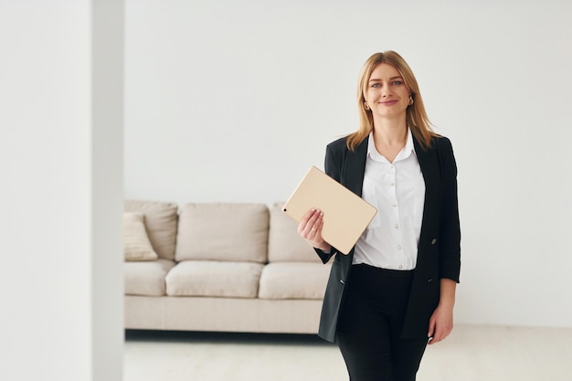 Woman in formal clothes standing near sofa indoors against white wall