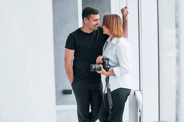 Женщина в формальной одежде и с камерой в руке стоит внутри пустой комнаты с мужчиной