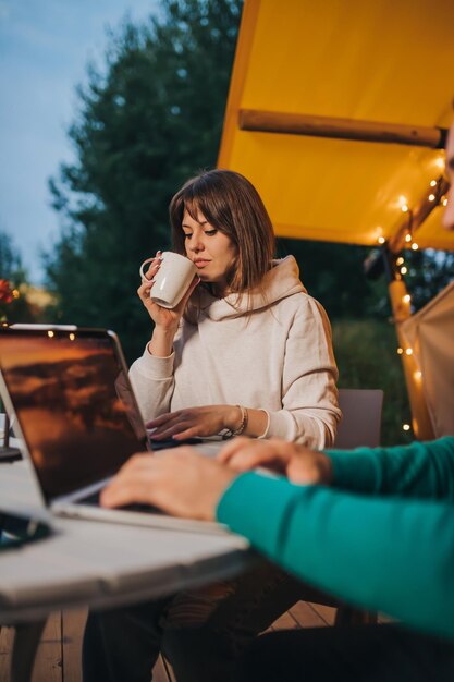 Woman freelancer drink coffee and working laptop near colleague\
on cozy glamping tent in summer evening luxury camping tent for\
outdoor holiday and vacation lifestyle concept