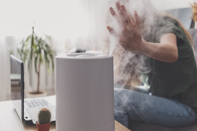 Woman freelancer uses a household humidifier in the workplace to maintain relative humidity and microclimate in the workplace of the home office with a laptop and documents.