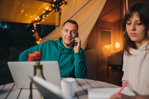 Woman freelancer working laptop and making notes while colleague talking phone sitting in cozy glamping tent in summer evening Luxury camping tent for outdoor holiday and vacation Lifestyle concept