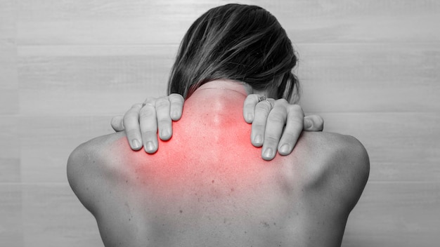 Woman from the back in black and white is palpating a red sore\
area on her back in the trapezius need for physical therapist or\
rehabilitation cerbic problem