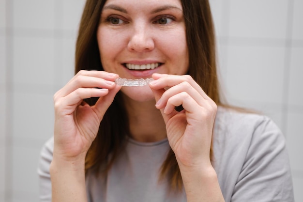 A woman in front of a mirror holding invisible plastic teeth aligners in hands Putting on braces