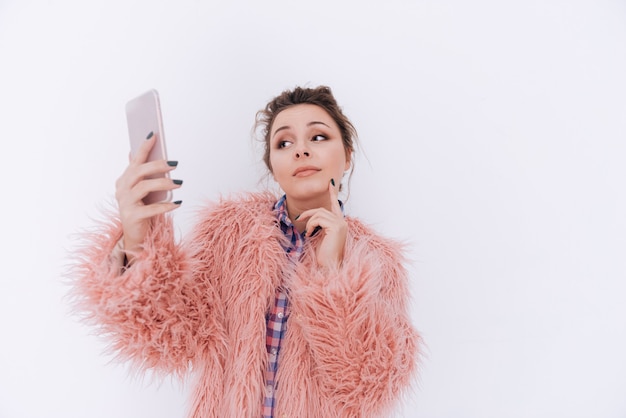 Woman in fur coat making selfie with hand near the face. Isolated gray wall