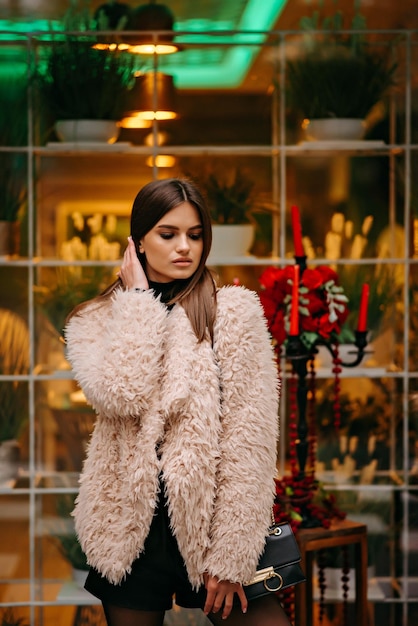 A woman in a fur coat stands in front of a window with a red candle behind her.