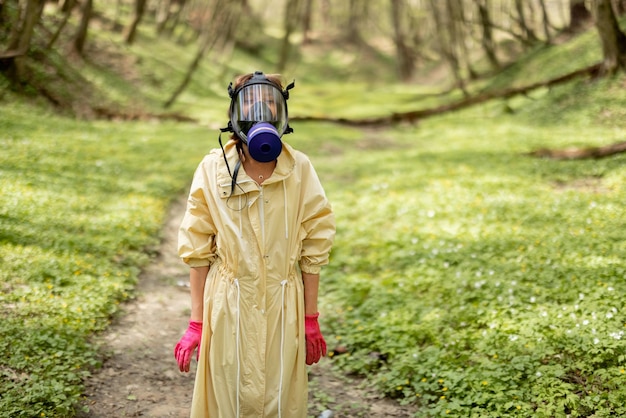 Foto donna in maschera antigas e indumenti protettivi che raccoglie rifiuti di plastica sparsi nei boschi