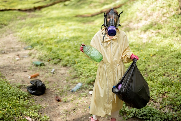 Foto donna in maschera antigas e indumenti protettivi che raccoglie rifiuti di plastica sparsi nei boschi