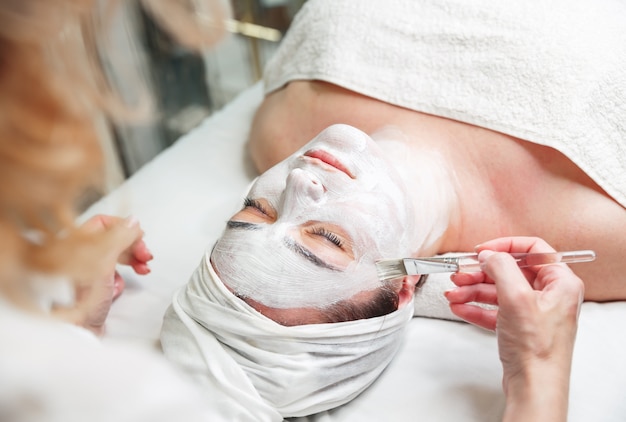 Woman gets face mask by beautician at spa salon. Cosmetician applying facial peeling mask.