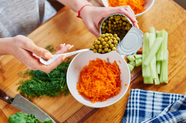 Woman gets a spoon out of a tin can with peas and puts in a bowl with the carrots standing on a wooden board