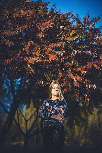 Woman girl long-haired blonde in a forest with trees with red leaves. She is happy confident in a photo shoot. Autumn park sunny warm day. Romantic concept.