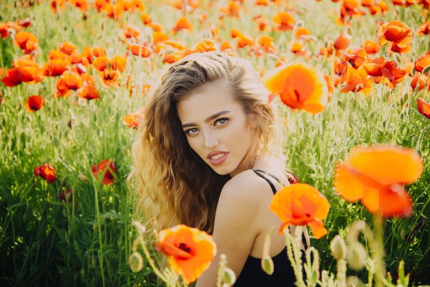 Woman girl with long curly hair hold flower in field of red poppy seed with green stem on natural ba...