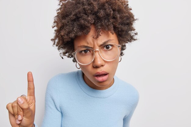 woman gives recommendation indicates with index finger above has dissatisfied expression wears optical glasses and blue jumper isolated on white