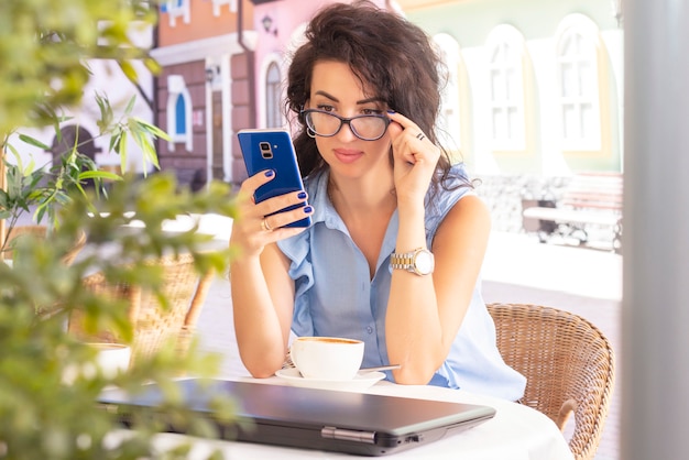カフェでノートパソコンと携帯電話でリモートで作業するガラスの女性。カフェでノートブックを使用して美しいブルネット。幸せな実業家を携帯電話で呼び出して撮影。フリーランス