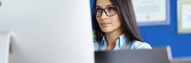 Woman in glasses and blue shirt sit at workplace and look at computer screen.