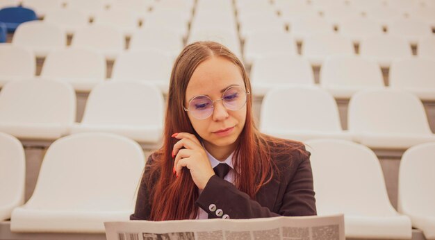 A woman in glasses and a business suit reads a newspaper while sitting at the stadium