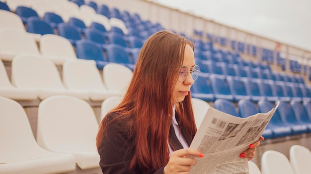 A woman in glasses and a business suit reads a newspaper while sitting at the stadium