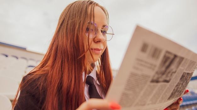 A woman in glasses and a business suit reads a newspaper while sitting at the stadium