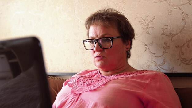 Photo a woman in glasses of a retirement age in a pink t-shirt works from home on a notebook.