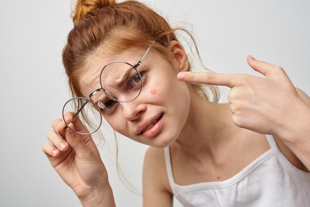 Woman in glasses shows a finger to a red pimple on her\
face.